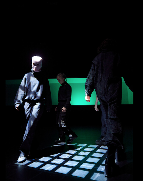 Szenografie und Kostüm von Lisa Behensky für ICARO – Figuren in dunklem Raum mit leuchtendem Bodenraster und maskierter Person im Vordergrund