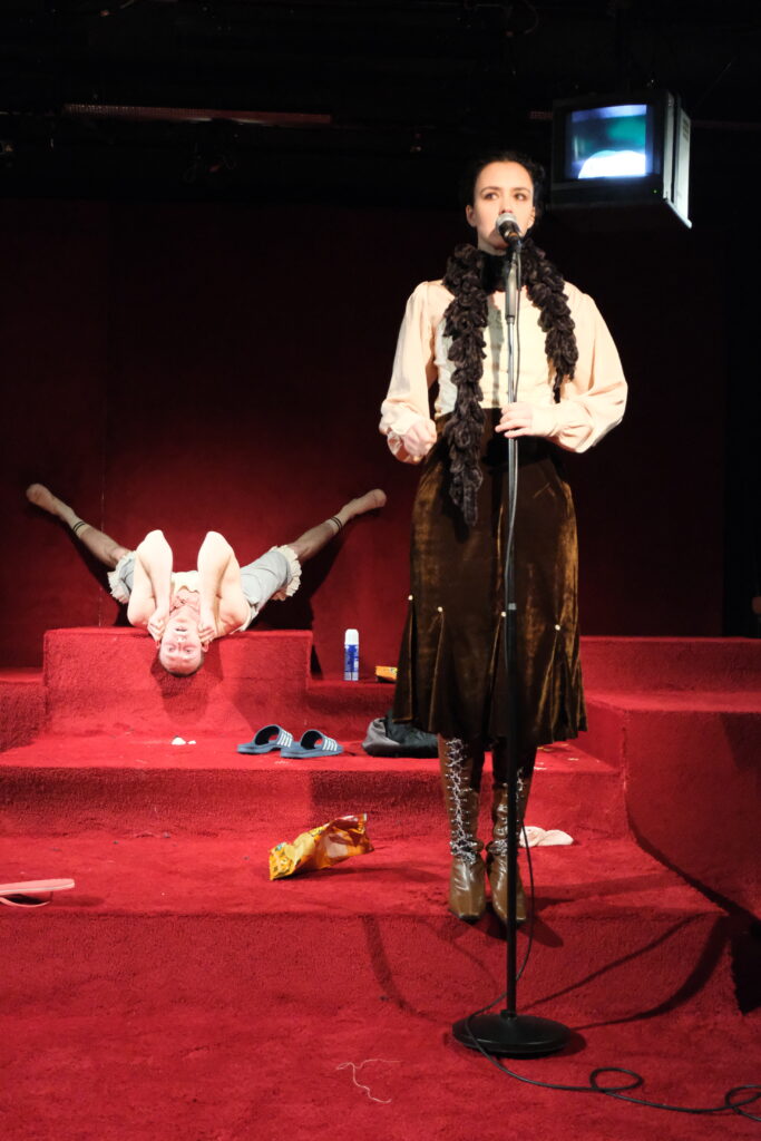 Szenografie und Kostüm von Lisa Behensky für Phaidras Liebe – zwei  Figuren vor roter Teppich-Wand mit Musikinstrumenten und starkem Hell-Dunkel-Kontrast