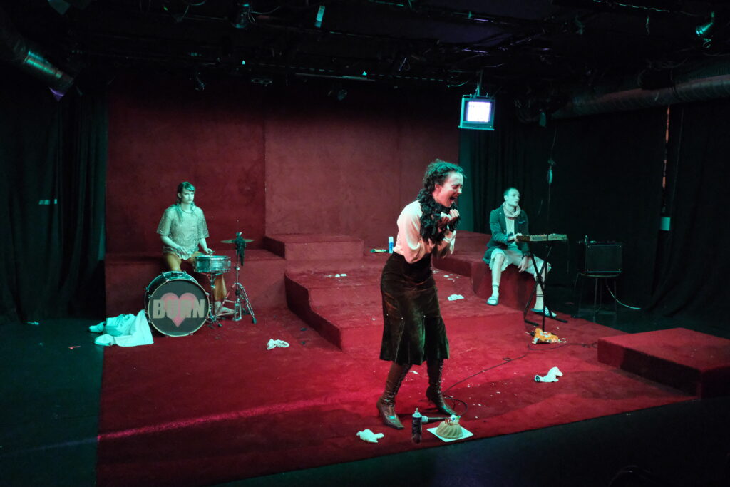 Szenografie und Kostüm von Lisa Behensky für Phaidras Liebe – drei isolierte Figuren in rotem Teppich Bühnenbild mit Musikinstrumenten und starkem Hell-Dunkel-Kontrast